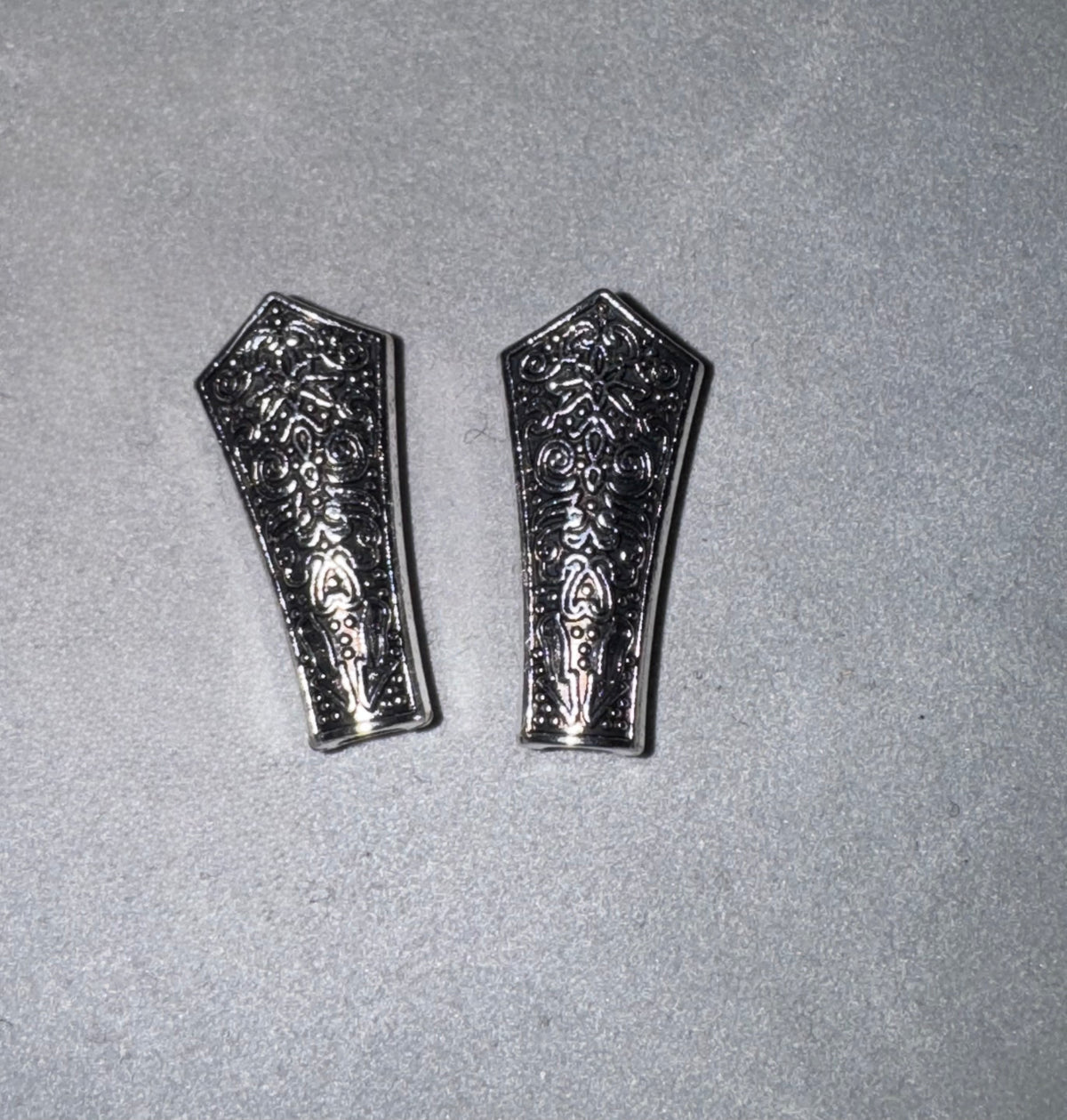 Two silver-toned metal decorative pieces with intricate floral engravings, displayed side-by-side on a textured gray background.