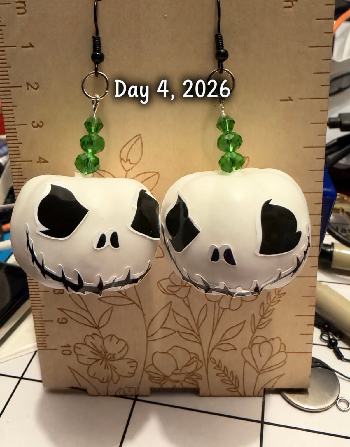 A pair of handmade jack-o'-lantern dangle earrings with green faceted beads, displayed on a wooden ruler with a date overlay reading 'Day 4, 2026'.