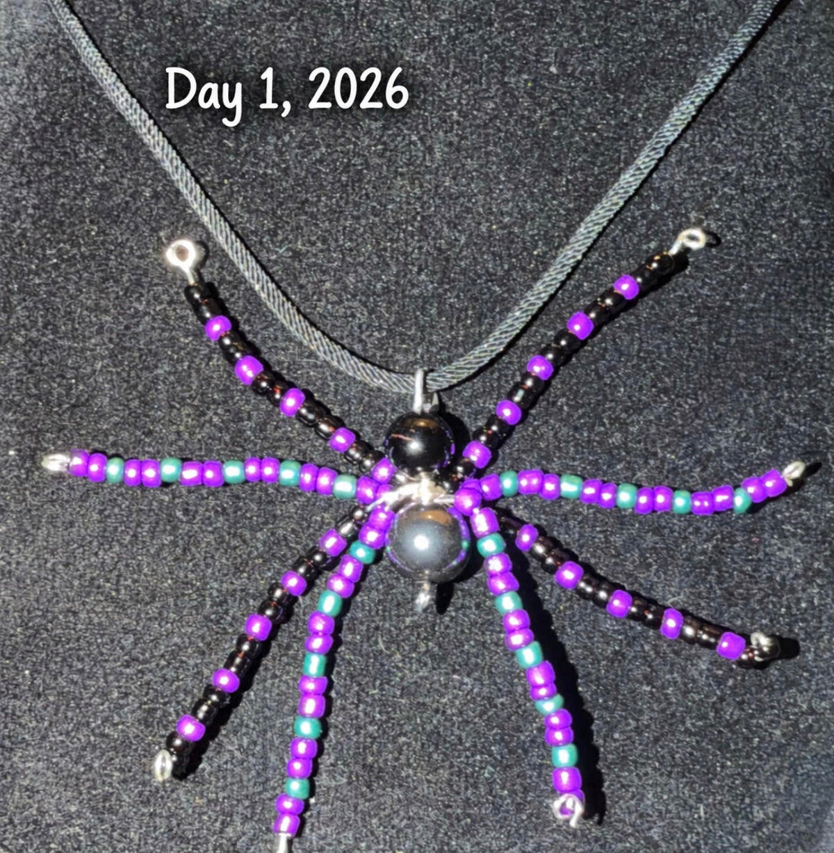 A handmade spider-shaped beaded pendant necklace on a dark gray velvet background, featuring purple, black, and mint green beads with a dark gray pearl center, with the text 'Day 1, 2026' written at the top.