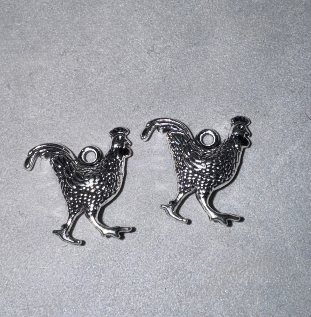 Two silver-toned metal rooster charms with textured scales and a loop at the top, displayed on a light gray textured background.