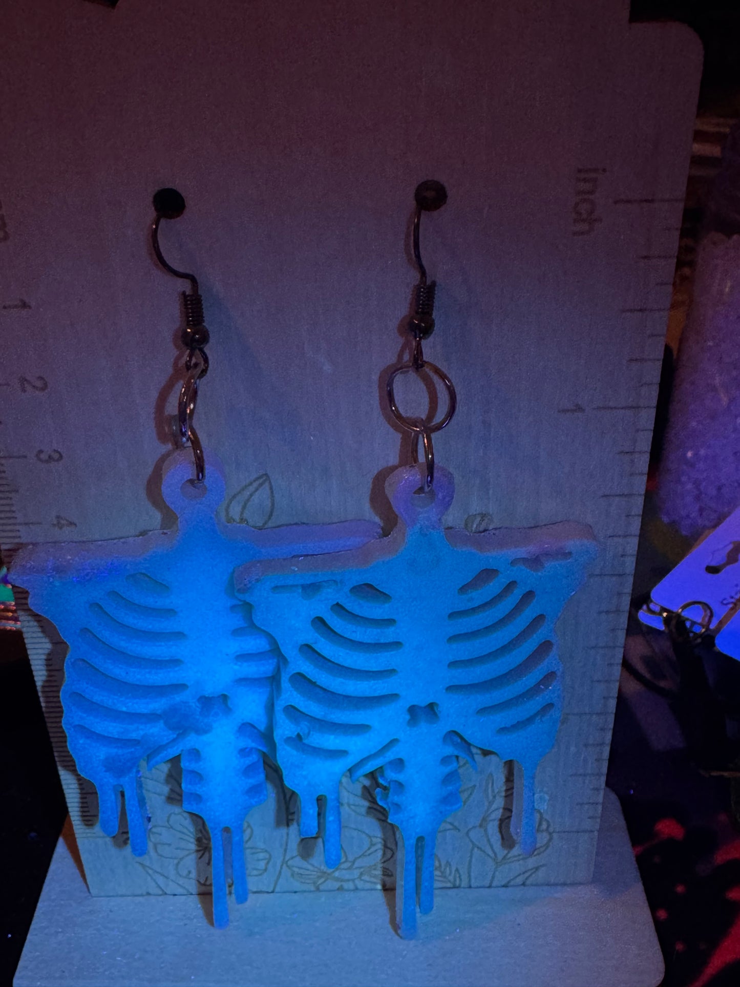 Blue skeleton-shaped earrings on a dark surface with a ruler for scale.