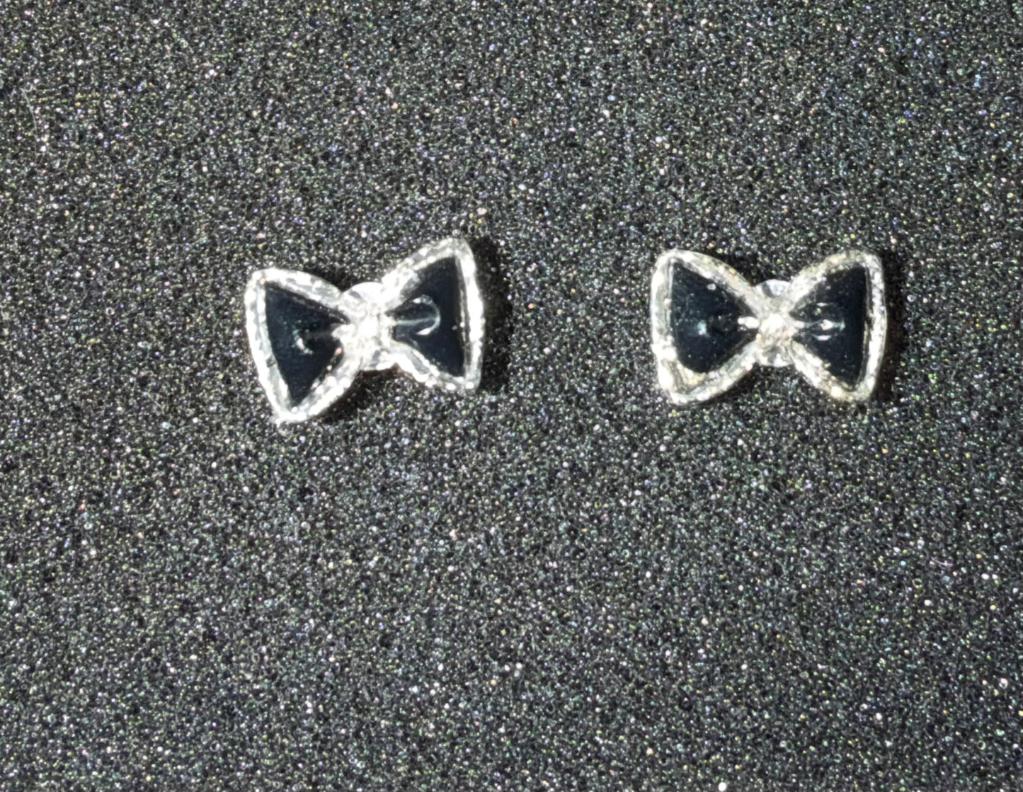 A pair of silver bow tie stud earrings with black enamel centers, displayed on a textured dark gray background.
