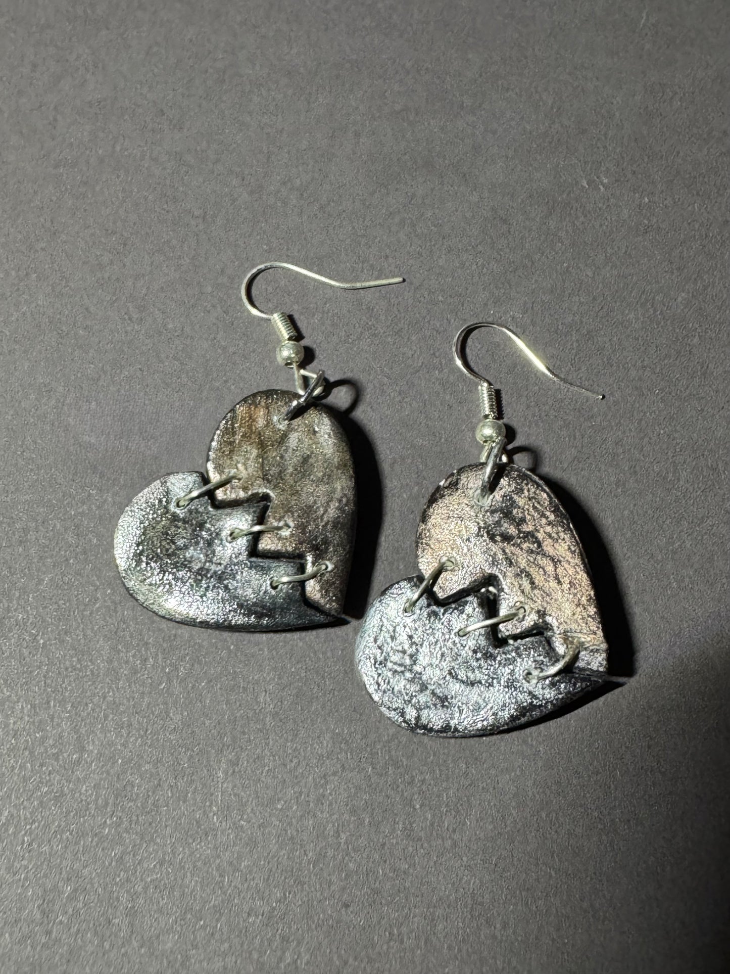 A pair of handmade heart-shaped dangle earrings with a distressed, metallic silver and bronze finish, featuring a broken-heart motif and silver fishhook wires, displayed on a dark gray background.