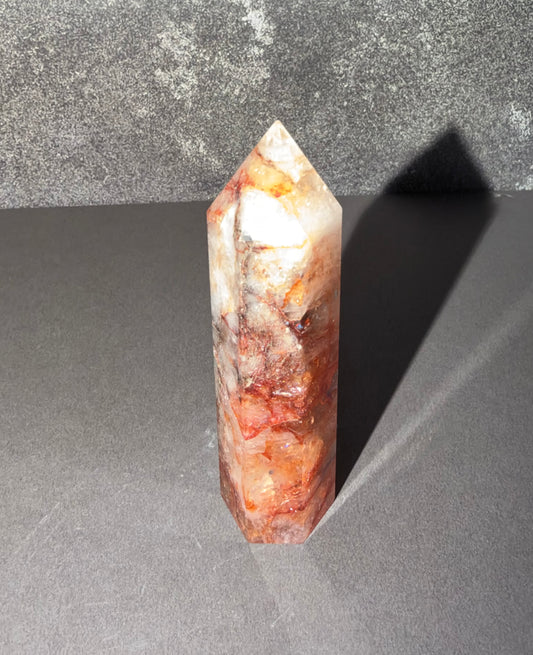 A polished crystal tower with natural marbled patterns in white, orange, and reddish-brown, standing upright on a dark gray surface against a textured gray background.