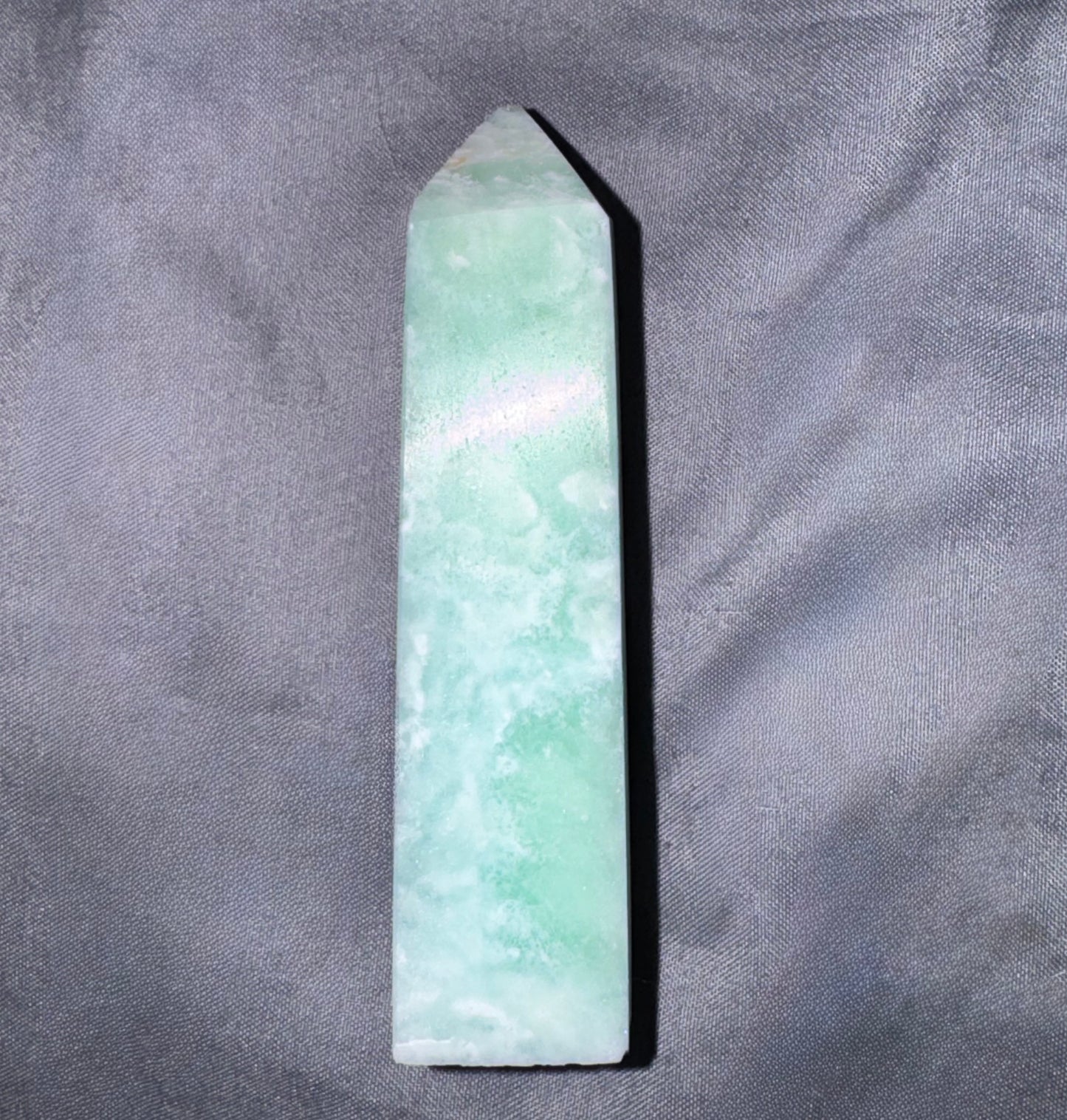 A pointed green jade crystal tower or wand, showing natural light green and white marbled patterns, resting on a textured gray fabric background.