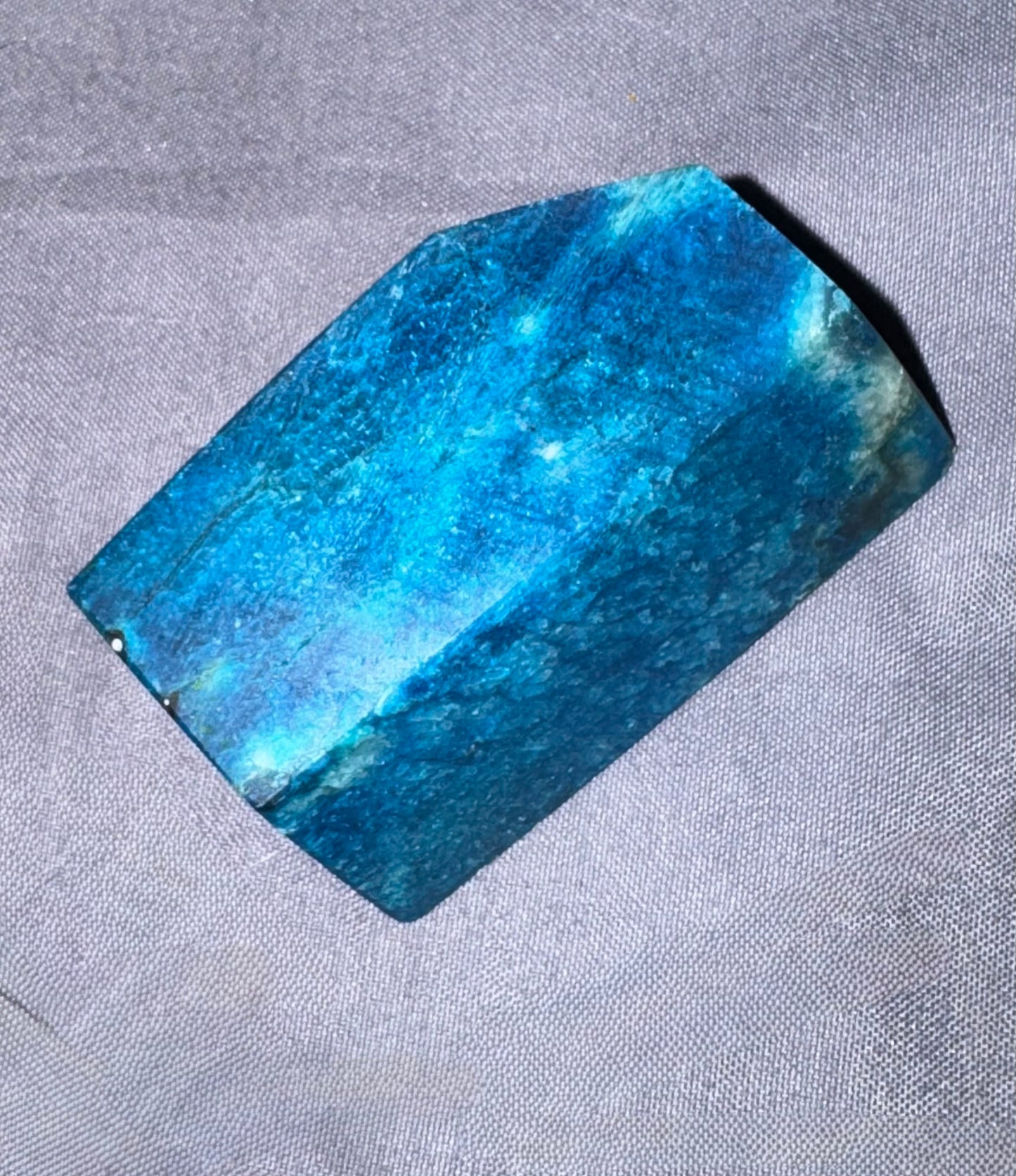 A polished blue stone or crystal with natural inclusions, photographed on a textured gray fabric background.
