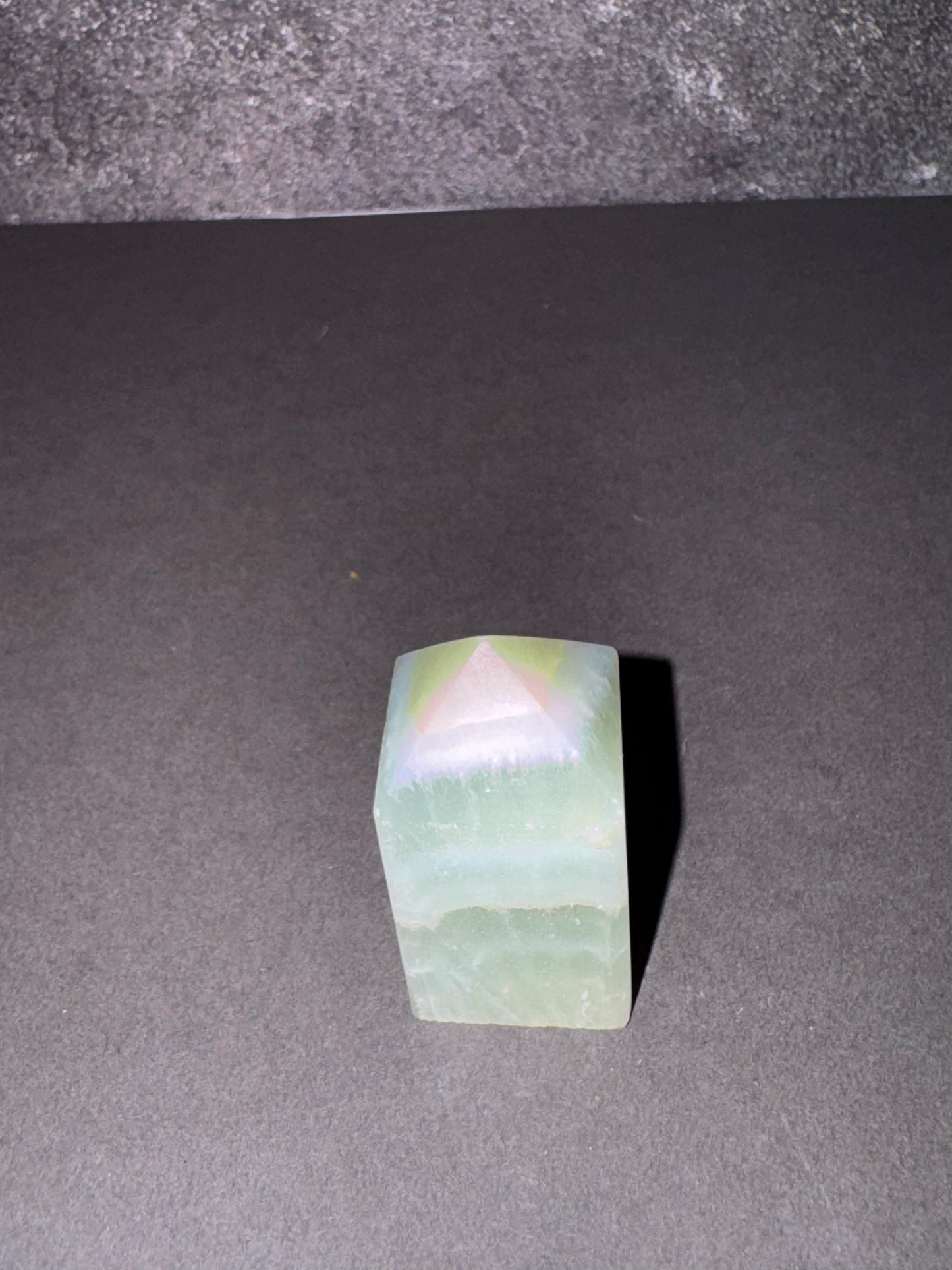 A small rectangular translucent green stone or crystal with a triangular pyramid cutout on top, standing on a dark gray surface against a dark gray background.