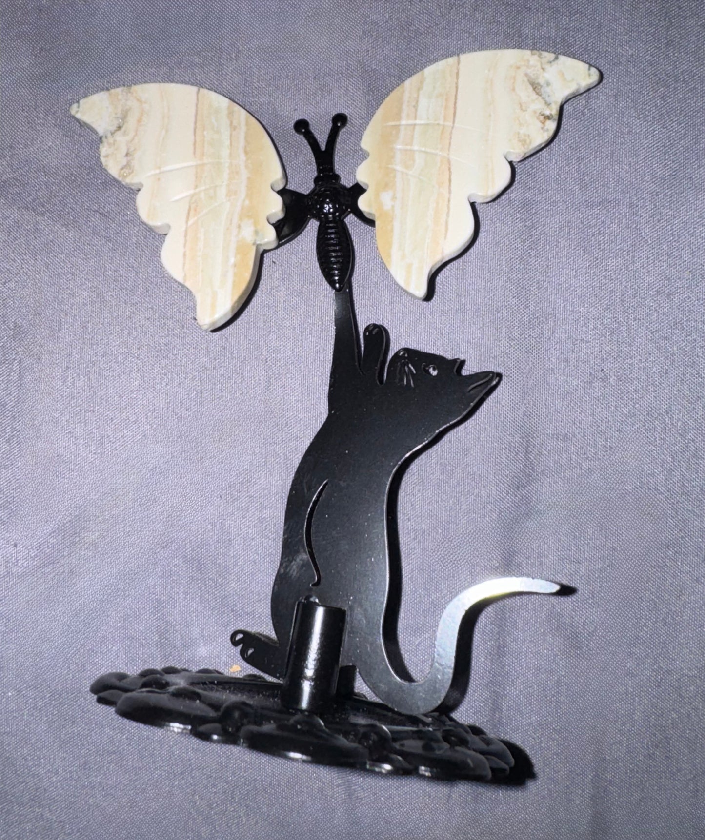 A decorative metal wall sculpture featuring a black cat silhouette reaching upward toward a butterfly with large cream-colored stone wings, mounted on a dark textured base against a gray fabric backdrop.