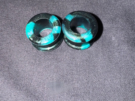 Two dark teal and black acrylic ear plugs with a distressed finish, displayed on a textured gray fabric background.