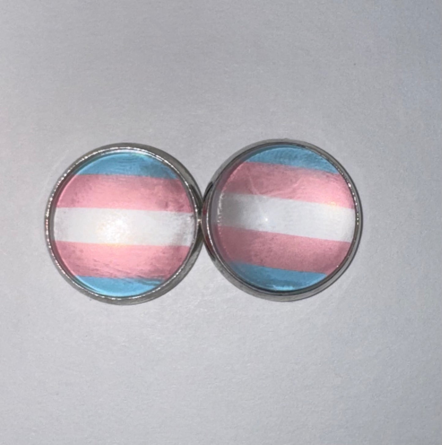 A pair of round stud earrings with a transgender pride flag design, featuring horizontal stripes of light blue, pink, and white under a clear dome, set in a silver-toned metal bezel, on a light gray background.
