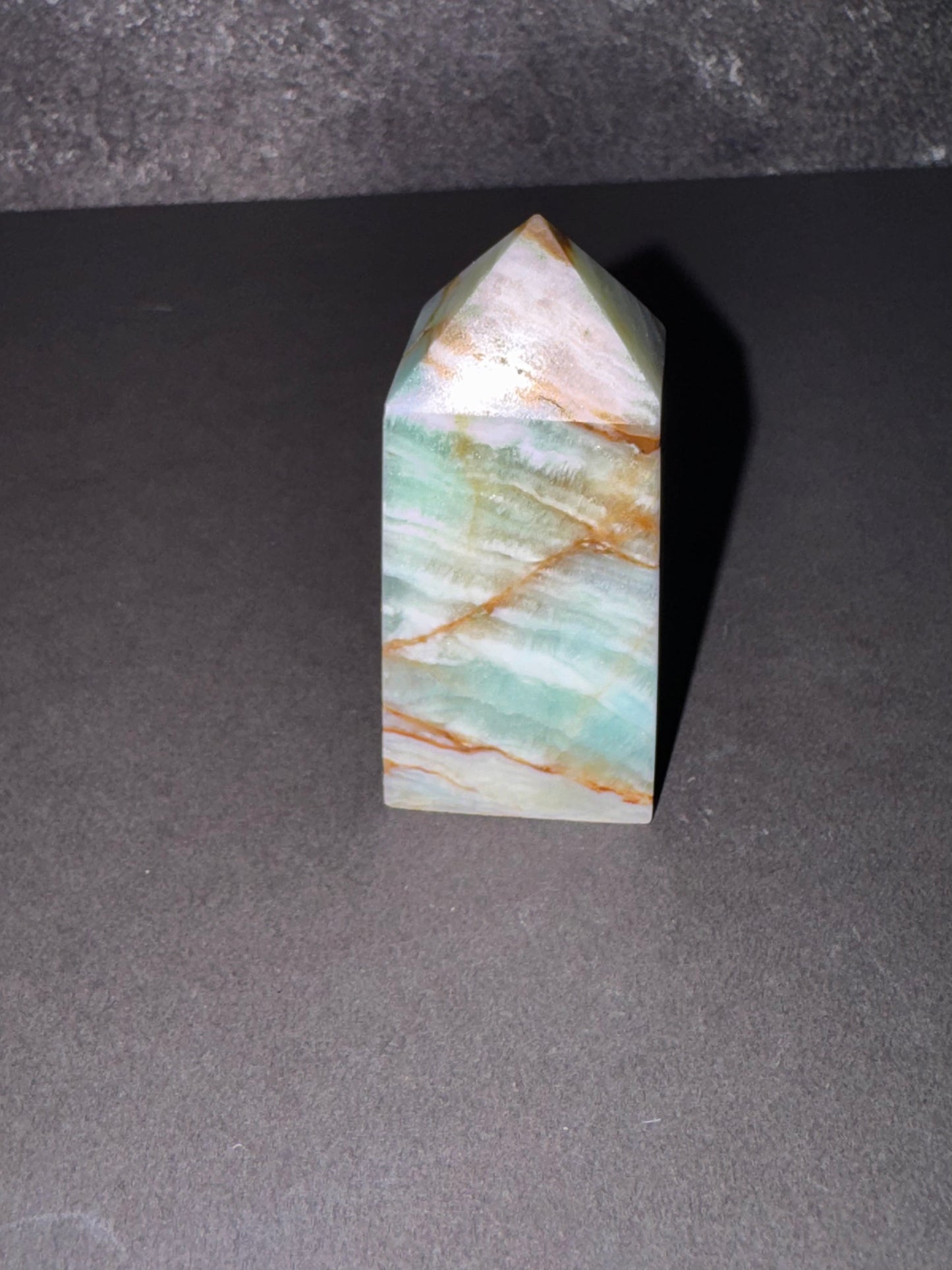 A natural quartz crystal pyramid with a pointed tip, featuring a marbled pattern of light green, white, and orange-brown veins, standing on a dark gray surface.