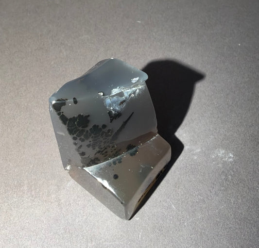 A polished natural crystal or mineral specimen with a translucent gray body and dark black inclusions, photographed on a neutral gray surface with a sharp shadow.