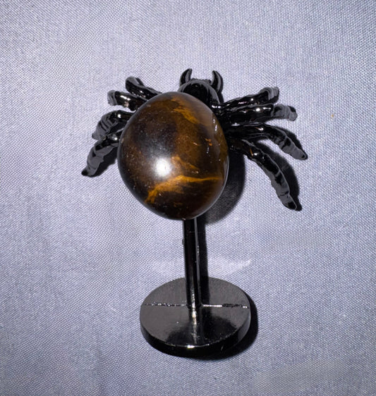 A decorative spider sculpture with glossy black legs and a polished brown tiger's eye sphere on a black stand, photographed against a textured light gray fabric background.