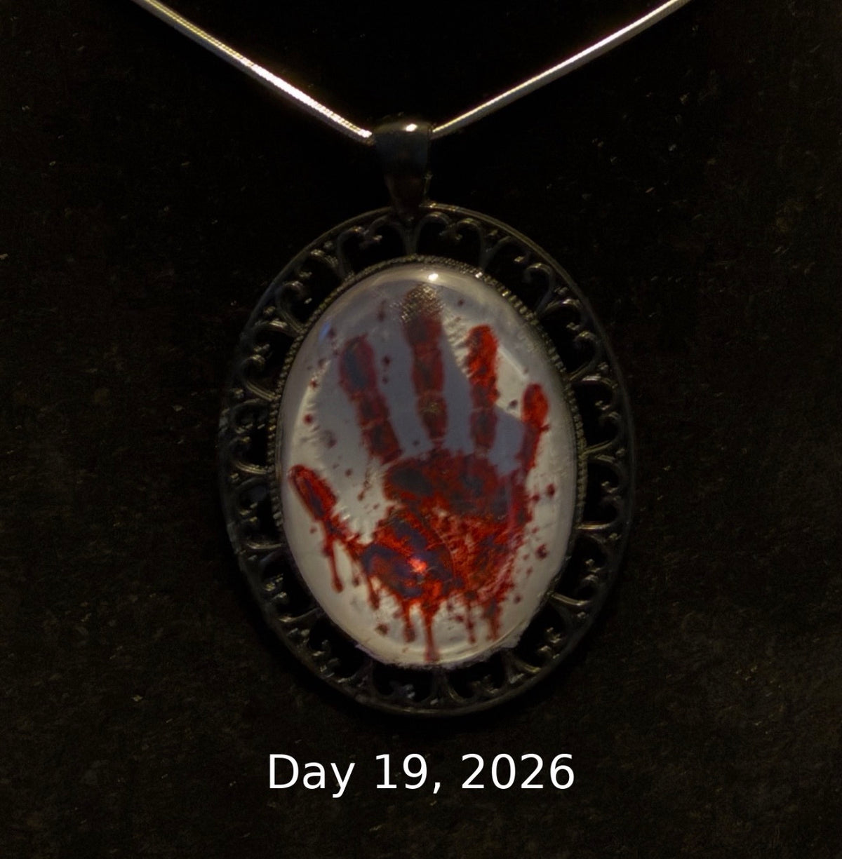 An oval pendant necklace with a black filigree-style border and a clear dome showing a white background with a red handprint design, suspended from a silver-toned chain against a dark background. White text at the bottom reads 'Day 19, 2026'.