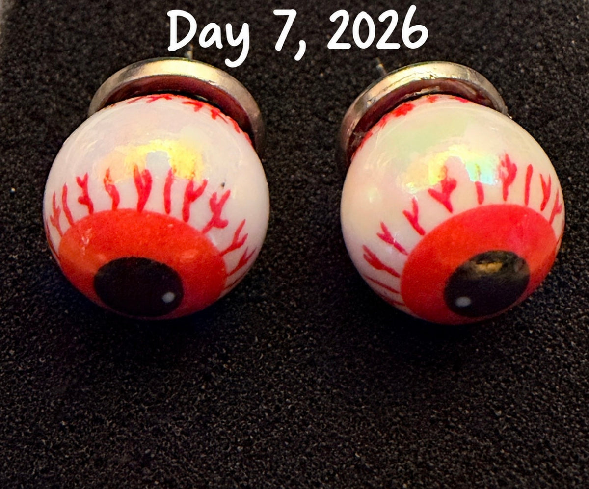 A pair of gothic-style eye stud earrings with white enamel, red blood-like veins, and realistic black pupils, displayed on a black textured background with a white text overlay reading 'Day 7, 2026'.
