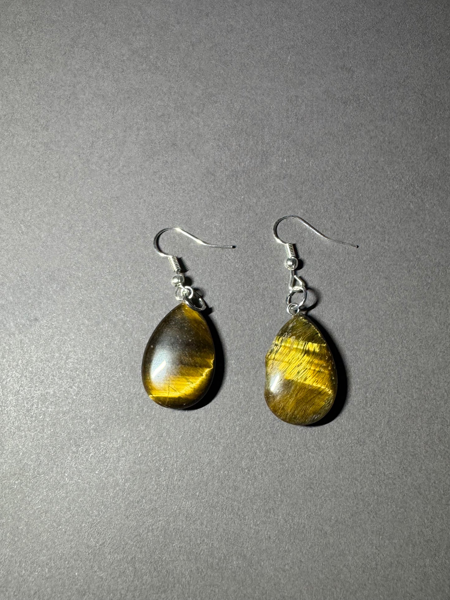 A pair of teardrop-shaped dangle earrings made from polished tiger's eye stones with silver fishhook wires, displayed on a textured gray background.