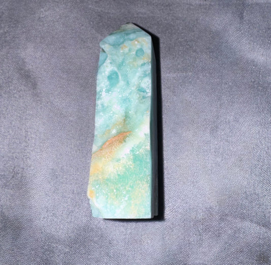 A polished natural crystal point with a marbled pattern of light blue, mint green, and soft orange, displayed on a textured gray fabric background.