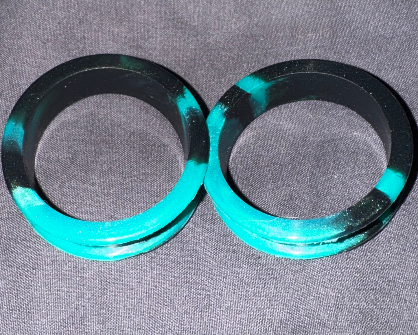 Two circular black rings with bright turquoise green painted sections, placed side by side on a gray textured fabric background.