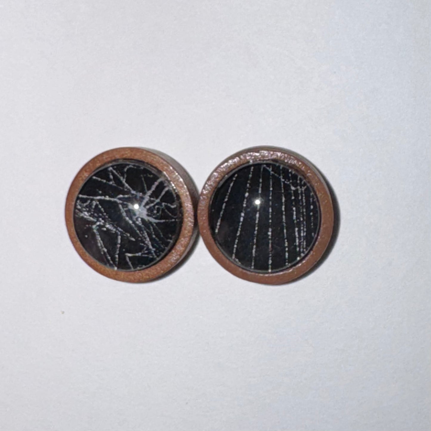Two round wooden cabochons with black and white cracked and vertical line patterns, set in a brown wood frame, on a light gray background.