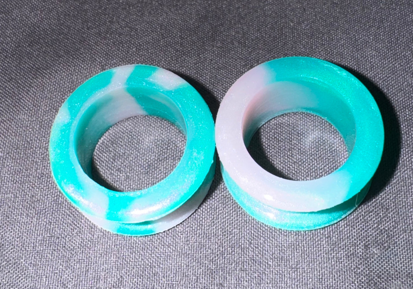 Two circular ring-shaped items with a marbled turquoise and white pattern, photographed on a textured gray fabric background.