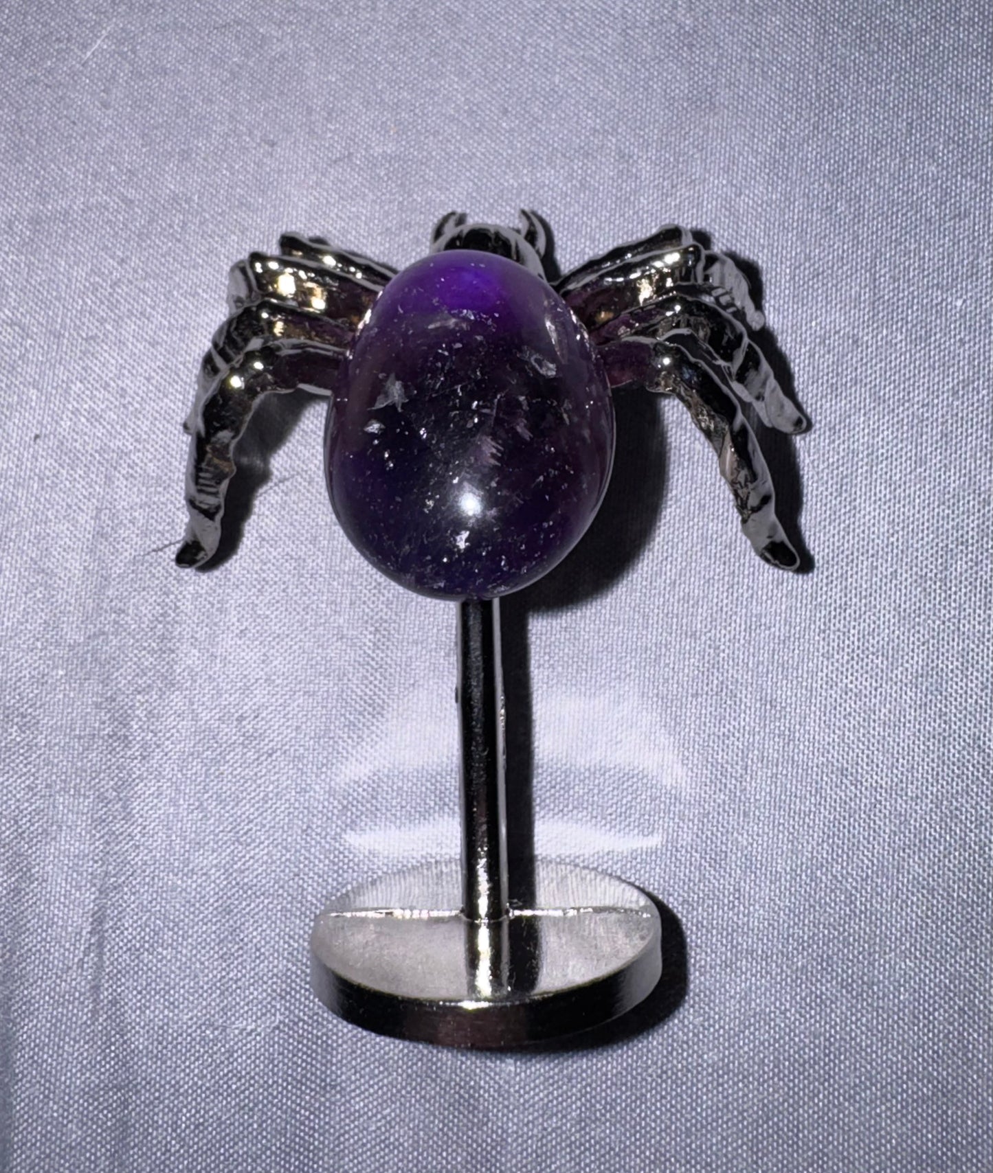 A spider with a polished purple amethyst egg center and metallic silver spider body and stand, displayed on a silver stand against a light gray fabric background.