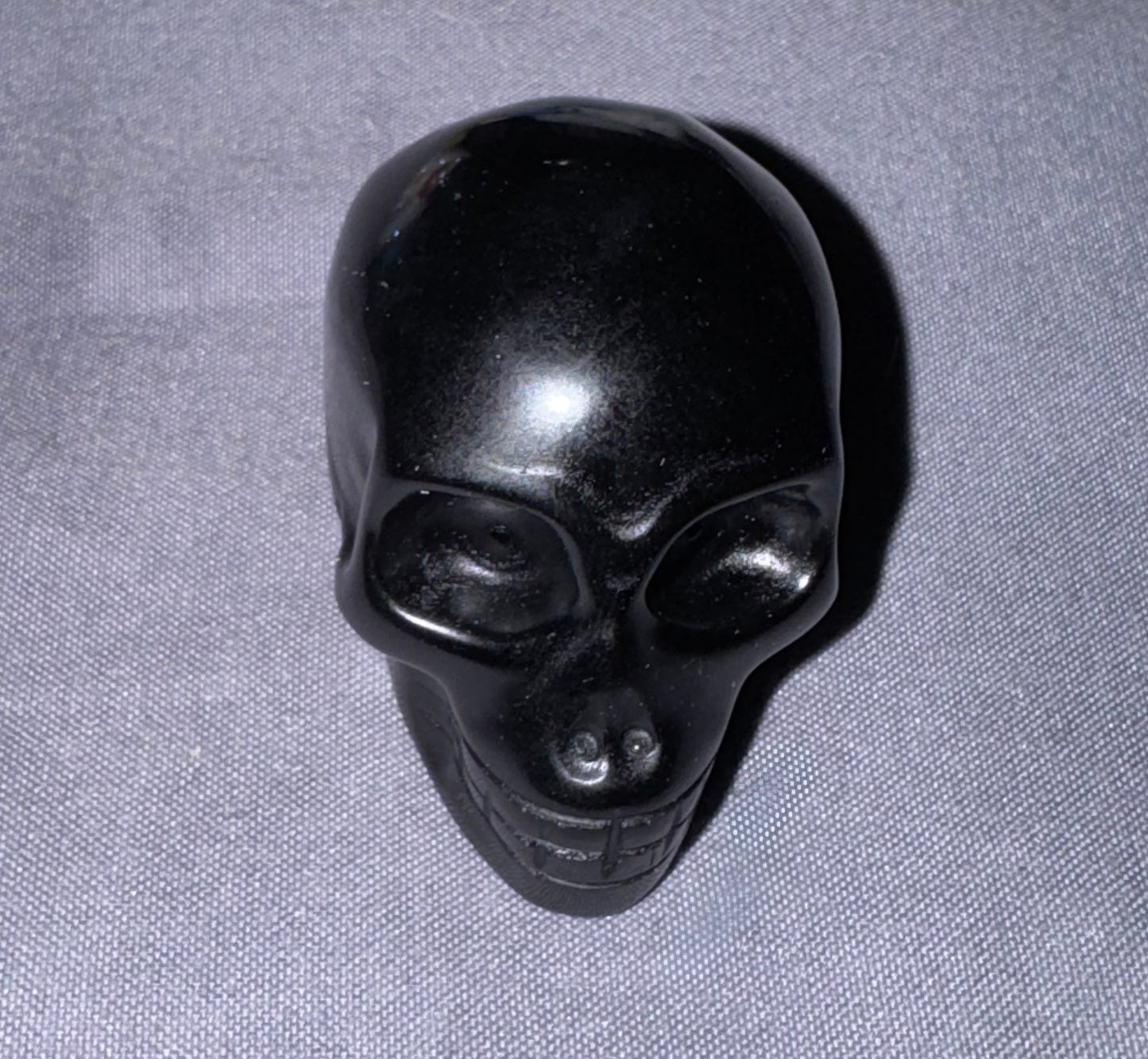 A glossy black skull-shaped object, possibly a decorative carving or small container, photographed on a textured gray fabric background.