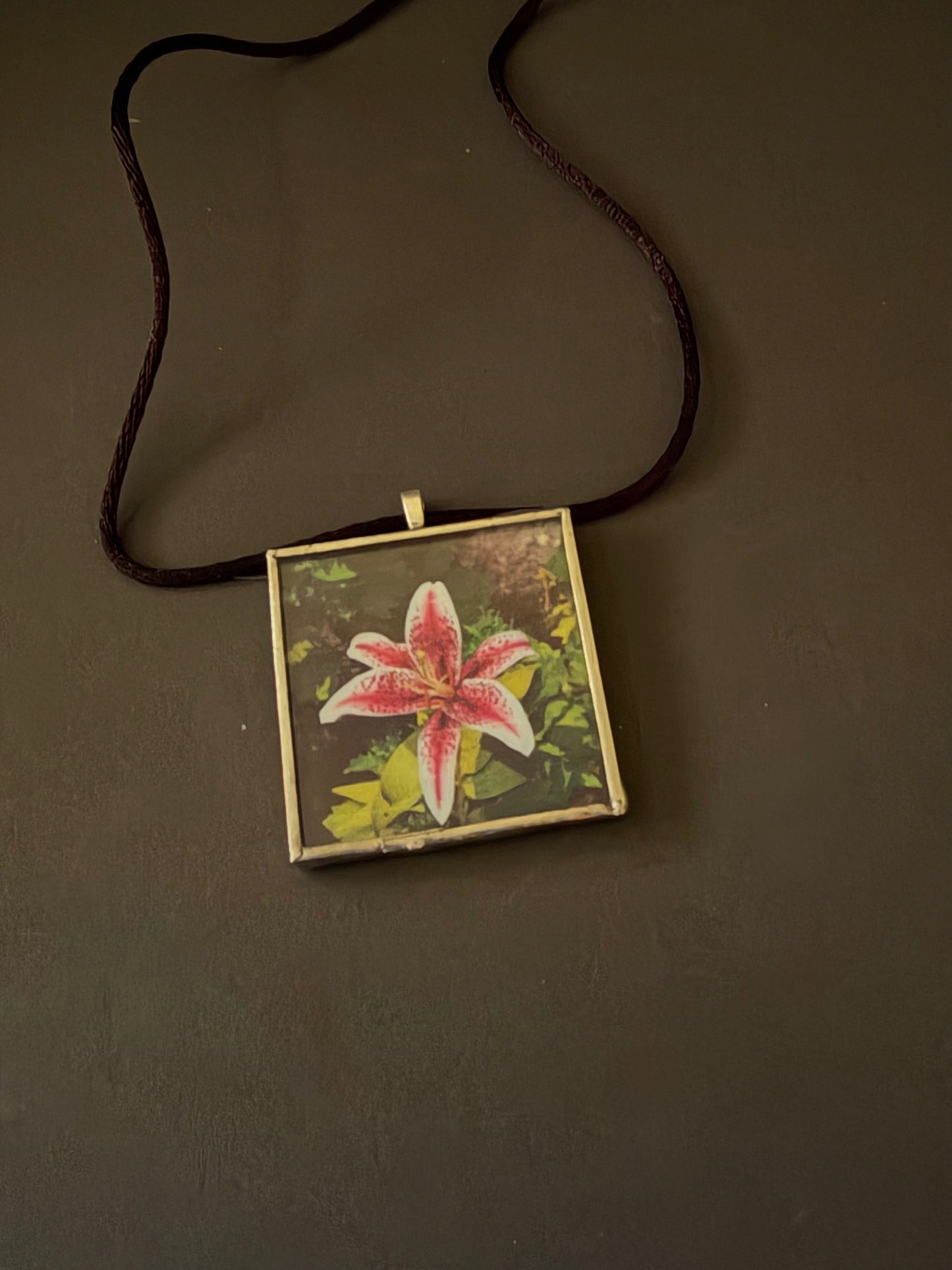 A square pendant necklace featuring a printed pink and white flower on a dark background, suspended from a dark brown cord against a dark brown surface.
