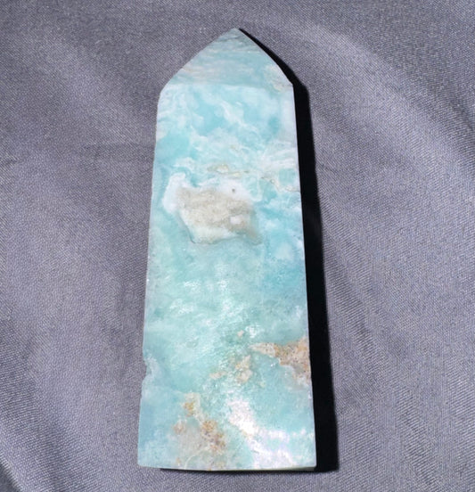 A polished crystal point, featuring natural light blue and white marbled patterns with small tan inclusions, displayed on a textured gray fabric background.