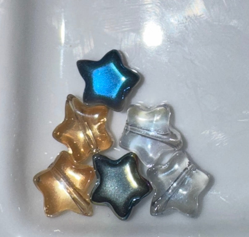 Five star-shaped beads in assorted colors, including metallic gold, iridescent blue, smoky gray, and clear, arranged on a white background.