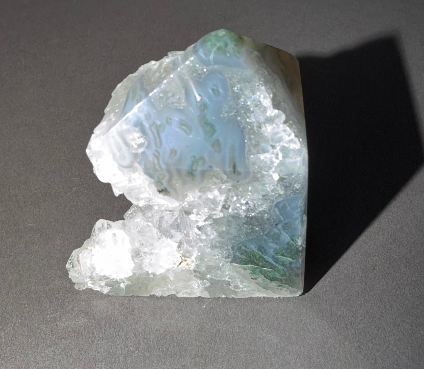 A raw agate crystal specimen with natural banded patterns in pale blue, white, and soft green, photographed on a dark gray surface with a sharp shadow.