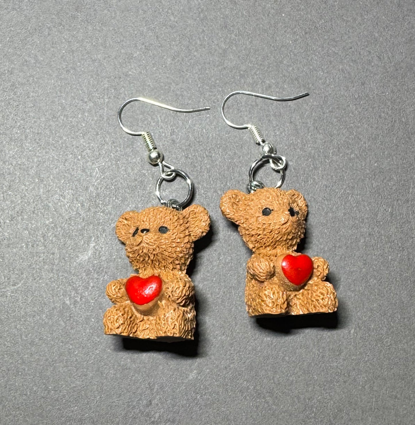 A pair of dangling earrings shaped like small textured brown teddy bears holding red hearts, suspended from silver fishhook wires on a gray background.
