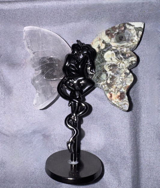 A winged figurine with a black metallic snake-wrapped body, a clear translucent left wing, and a mottled gray-green right wing, displayed on a round black base against a gray fabric backdrop.
