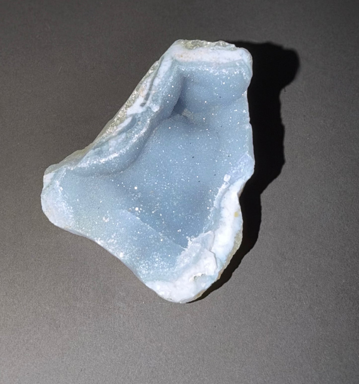 A raw, unpolished geode specimen with a light blue interior and white crystalline outer layers, photographed on a neutral gray background with a sharp shadow.