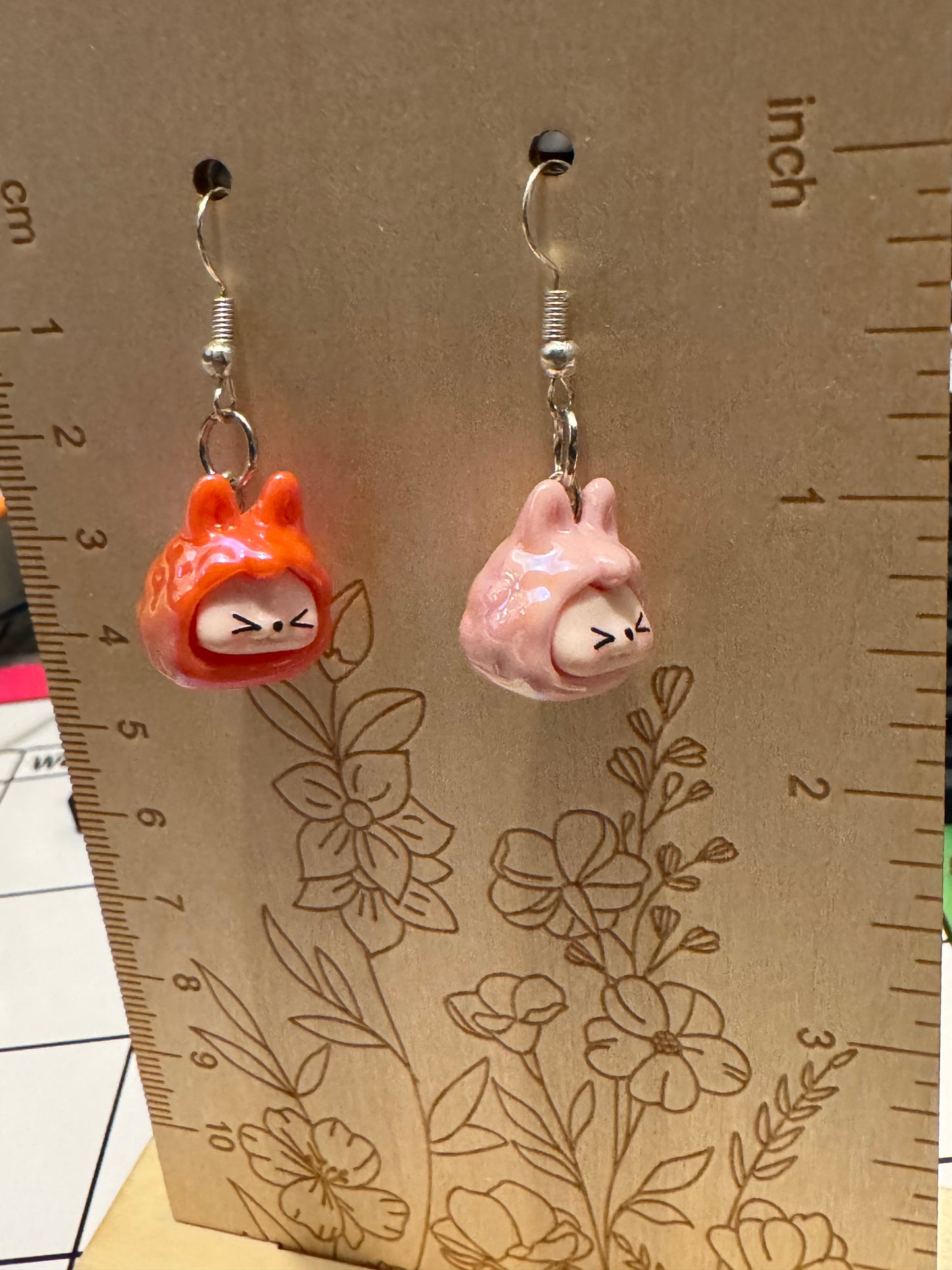 Two small dangling earrings displayed on a wooden measuring card. One earring has an orange-red hooded figure with a white face, and the other has a pink rabbit-like figure with a white face.