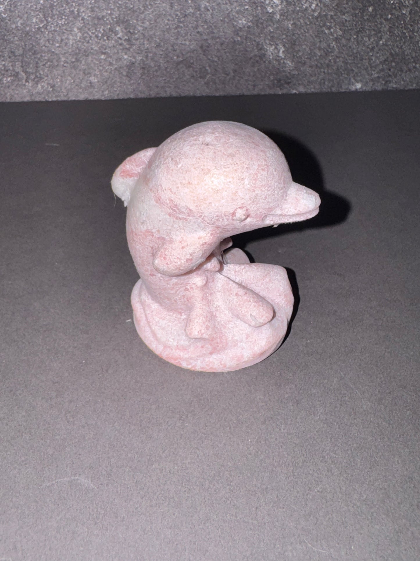 A small pink stone figurine of a seated bird, likely a dove or pigeon, photographed on a dark gray surface against a textured gray background.
