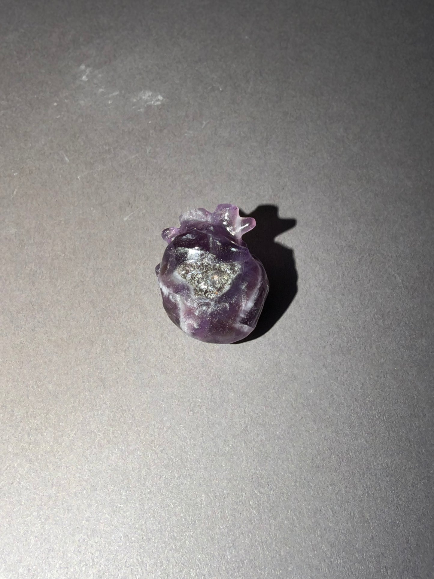 A small carved amethyst anatomical Heart with a clear crystal center, resting on a gray surface with a soft shadow.
