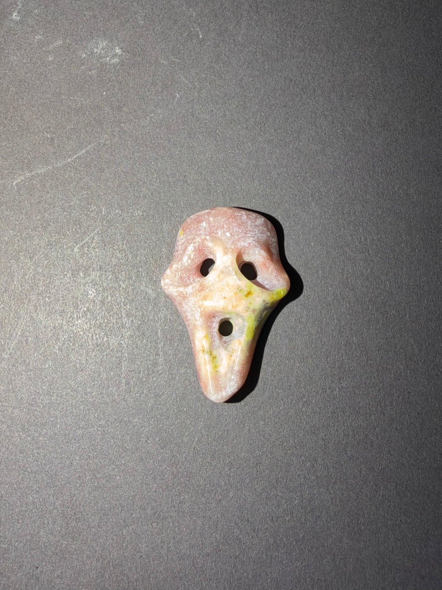 A small carved stone mask with two eye holes and a central opening, shown on a dark gray textured background.