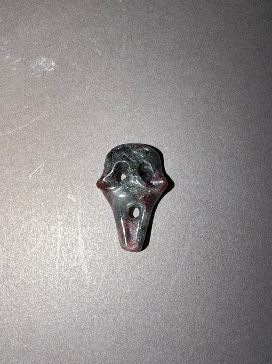 A small carved stone mask with a dark gray and reddish-brown marbled pattern, photographed on a plain gray surface.