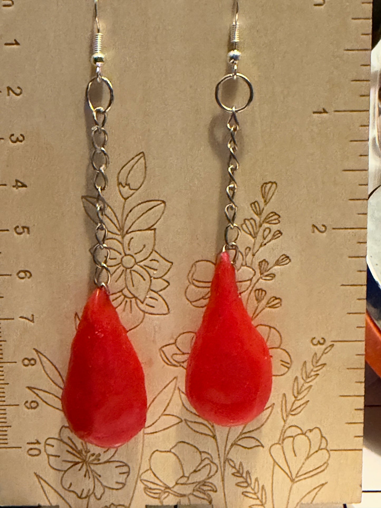 A pair of handmade dangle earrings with bright red teardrop-shaped beads hanging from silver-toned chains, displayed against a beige floral ruler background.