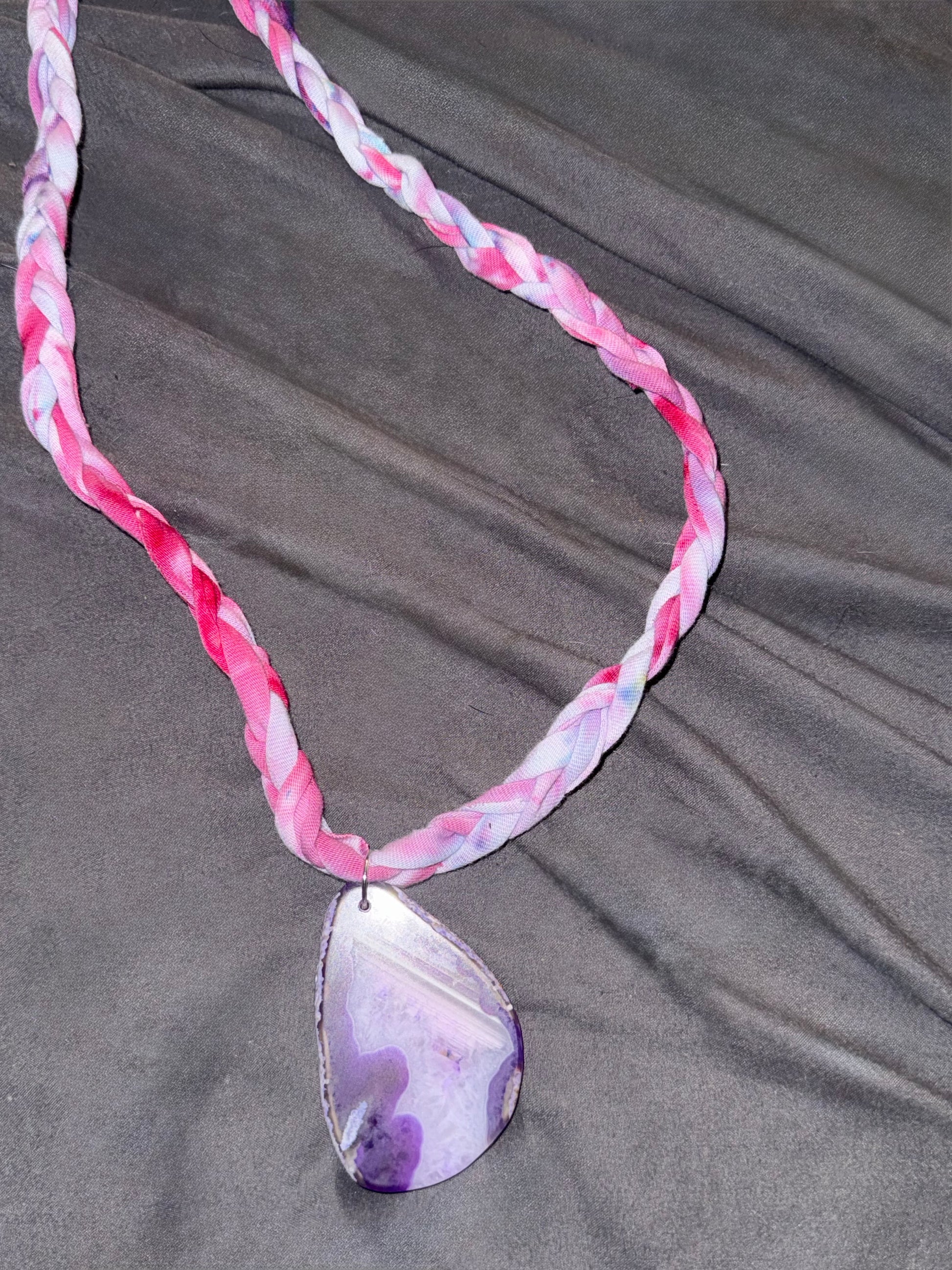A braided fabric necklace in shades of pink, white, and purple, featuring a polished teardrop-shaped pendant made from a purple and white agate slice, displayed on a dark gray fabric background.