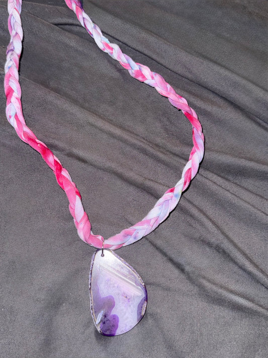 A braided fabric necklace in shades of pink, white, and purple, featuring a polished teardrop-shaped pendant made from a purple and white agate slice, displayed on a dark gray fabric background.