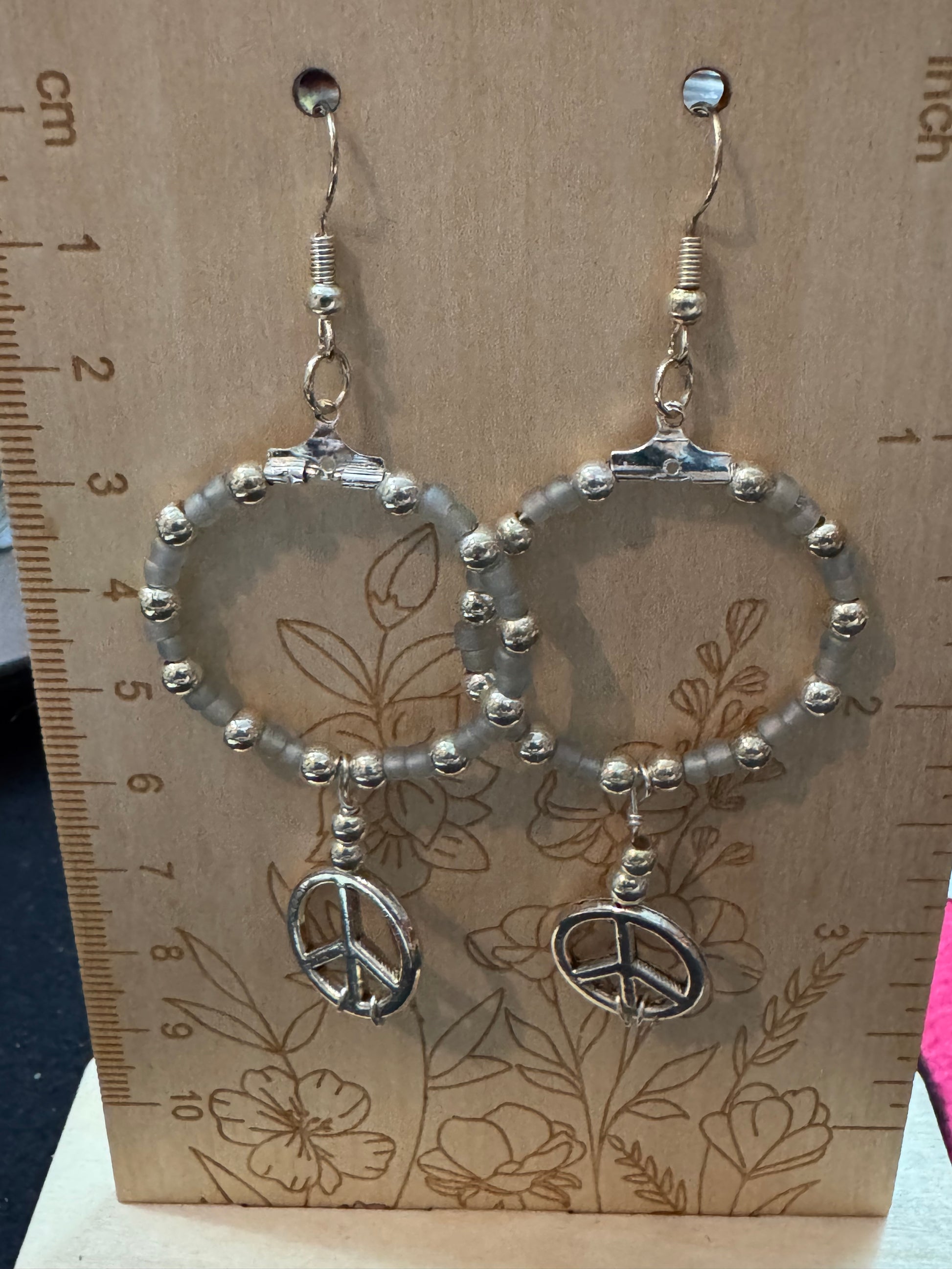 Silver earrings with peace sign charms on a floral-patterned card with a ruler for scale.