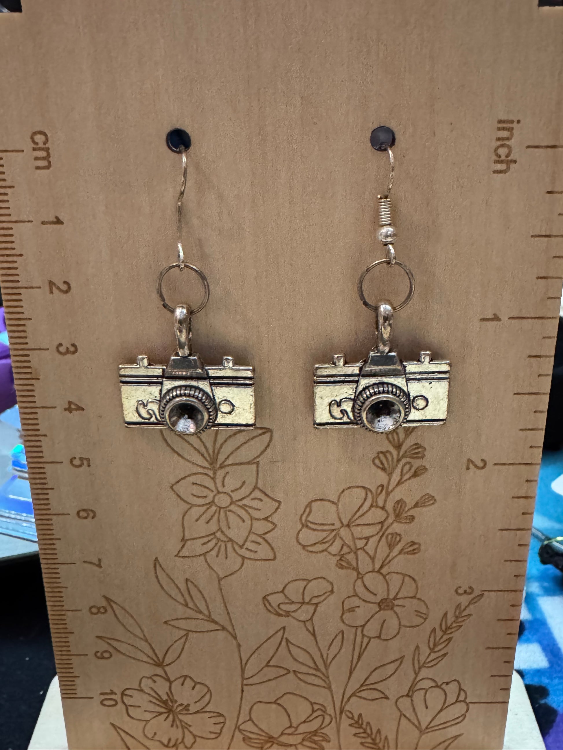 Camera-shaped earrings on a wooden board with floral patterns and measurements.