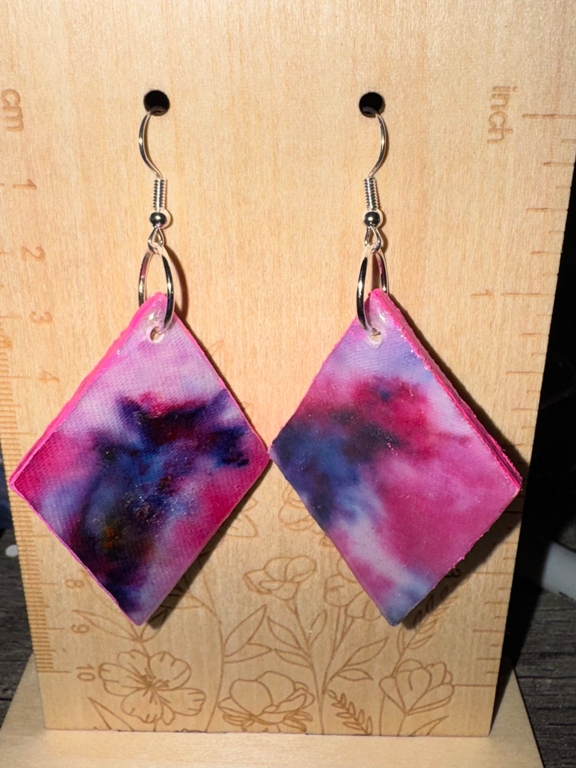 Pair of pink and purple abstract patterned earrings on a wooden stand with measurements.