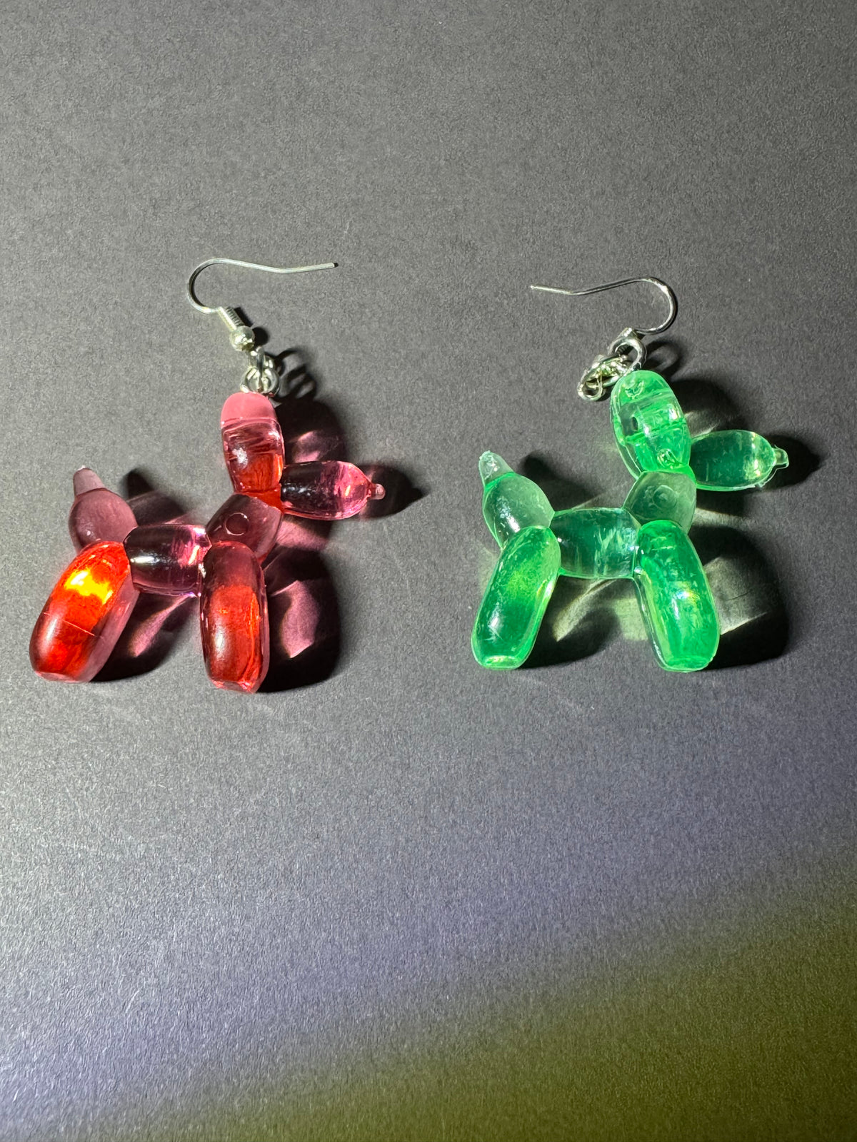 A pair of balloon dog dangle earrings, one translucent pink with red highlights and the other translucent green, shown on a dark gray background.