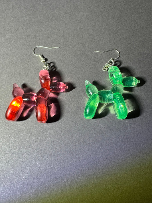 A pair of balloon dog dangle earrings, one translucent pink with red highlights and the other translucent green, shown on a dark gray background.