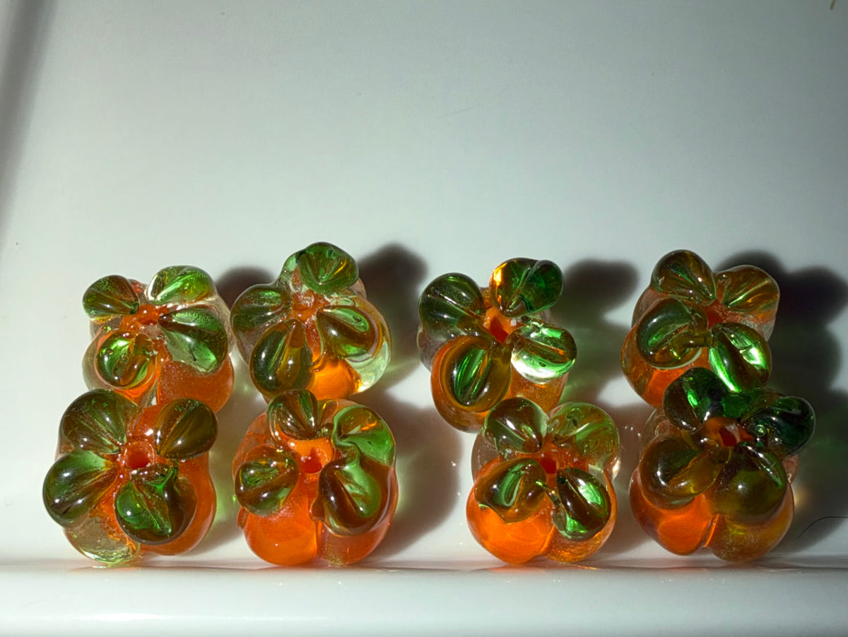 Six small translucent glass pumpkin-shaped beads with green petals and orange centers, arranged on a white surface under direct light.