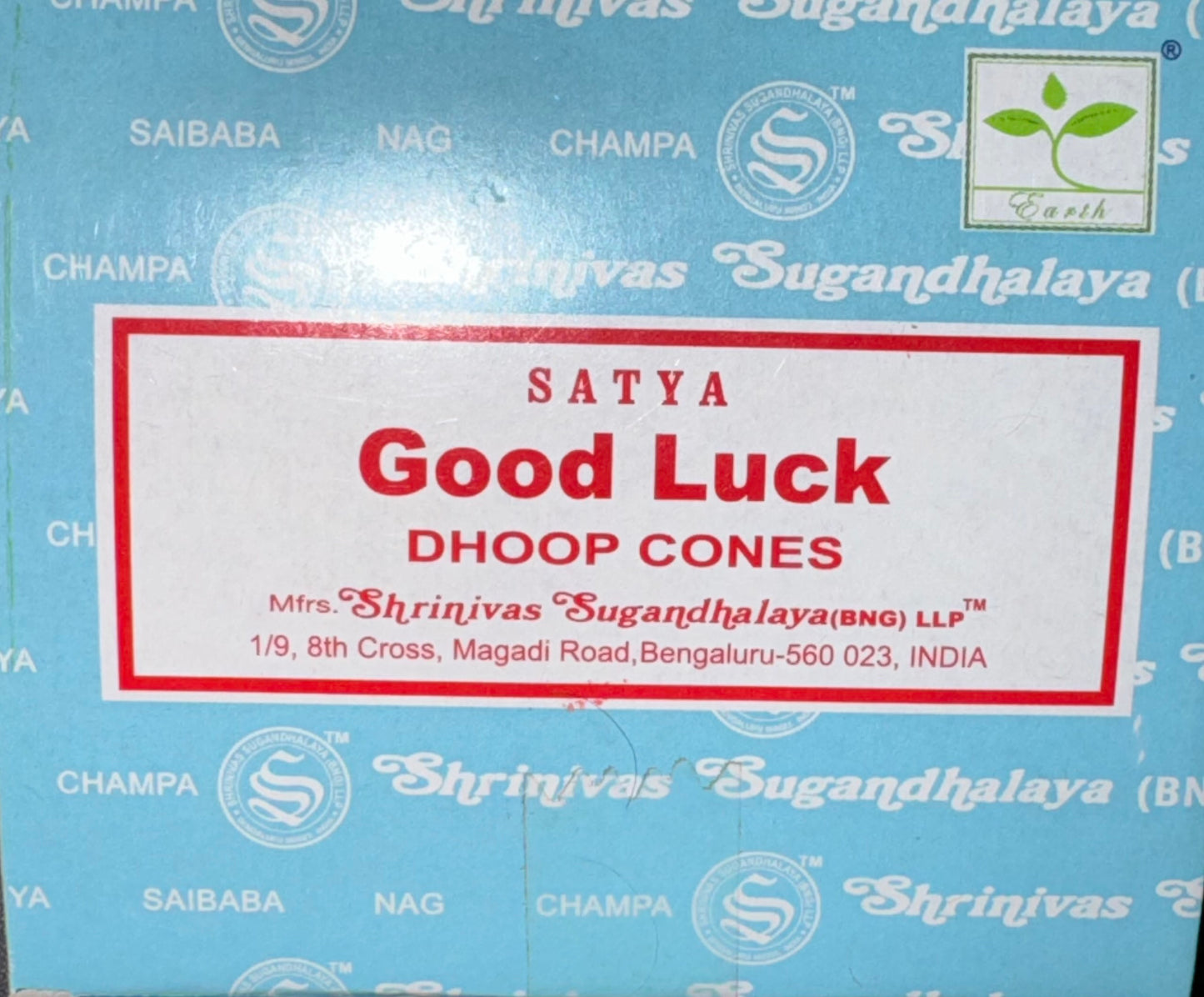 A close-up of a blue box of Satya Good Luck Dhoop Cones, featuring a white label with red text and border. The box has a repeating pattern of other product names like Champa, Nag, and Saibaba, along with the Surya logo.