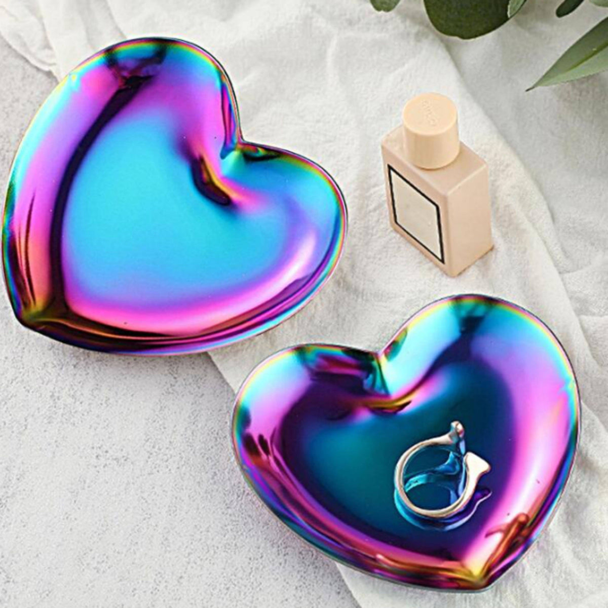 Two heart-shaped iridescent trays with a rainbow holographic finish, one empty and one holding a silver ring, styled on a white fabric surface with a small perfume bottle and green leaves in the background.