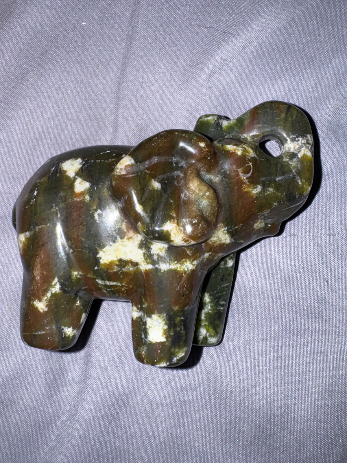 A carved elephant figurine made of polished stone with natural brown, olive green, and cream marbling, resting on a gray fabric background.